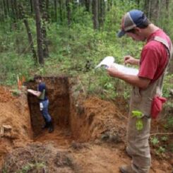 man in overalls and a red shirt observing a student studying layers of soil in a hole of earth 